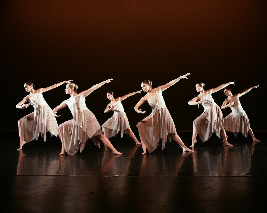 Stage image from a Spring Dance Concert showing a group of dancers arranged in a horizontal line, each extending one arm outward in synchronized motion. The performers wear soft, flowing dresses in light neutral tones that move with the choreography. The stage is dark with warm amber lighting that creates a gradient backdrop and highlights the shapes and lines of the movement.