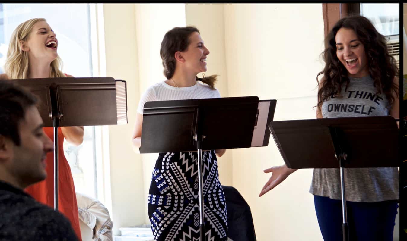 Rehearsal scene from a Stockton Theatre Project production, with several performers standing and singing beside black music stands. The group is positioned in a bright indoor space with light-colored walls and natural light, suggesting a casual performance or practice environment.