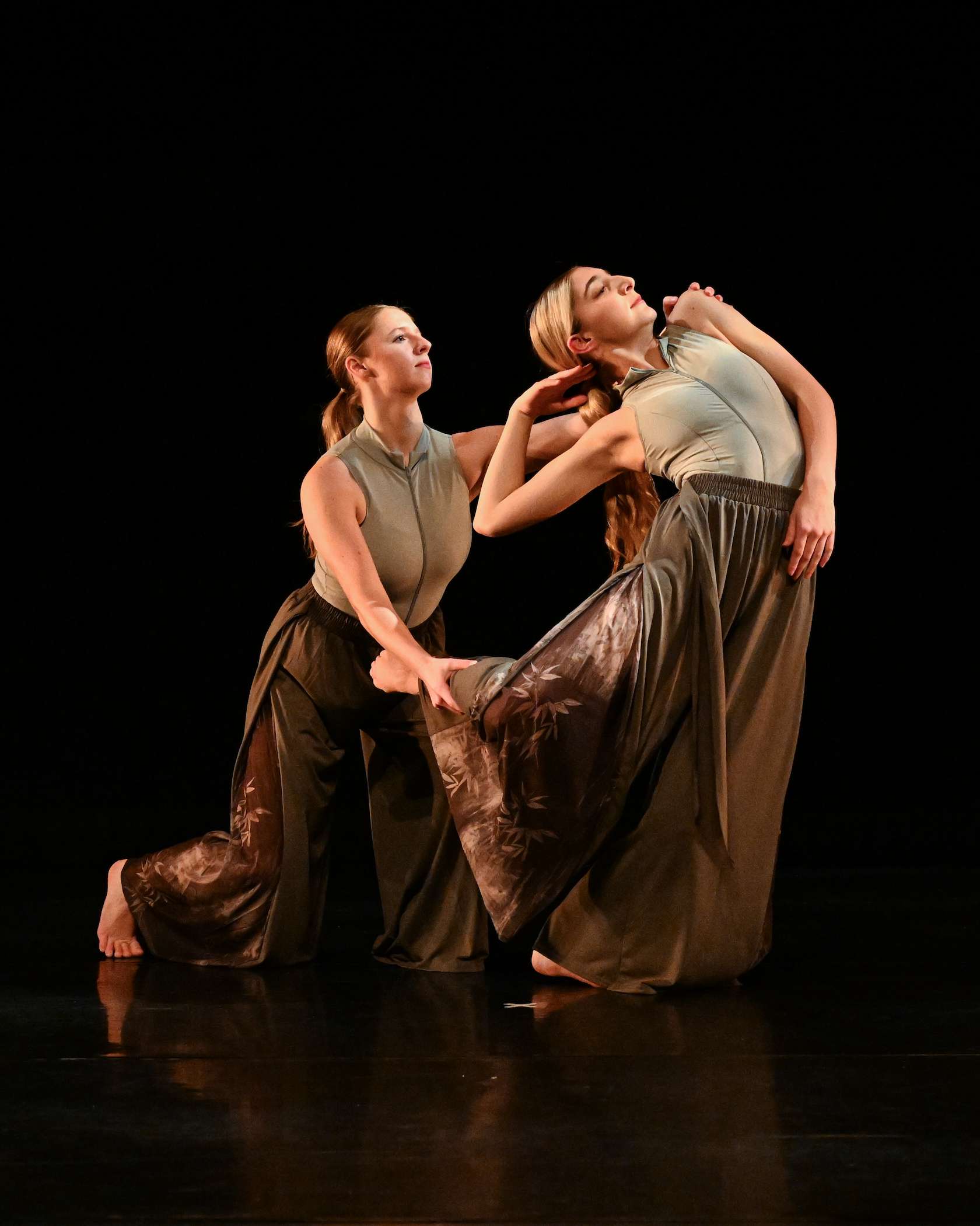 Stage image from a Fall Choreography Project featuring two dancers in coordinated, flowing costumes in muted green and brown tones. One dancer kneels low with an arm extended, holding the fabric of the other dancer’s skirt, while the second dancer arches backward in a sculptural pose. The performance takes place on a dark stage with focused lighting highlighting their movement and the sheen of the fabric.