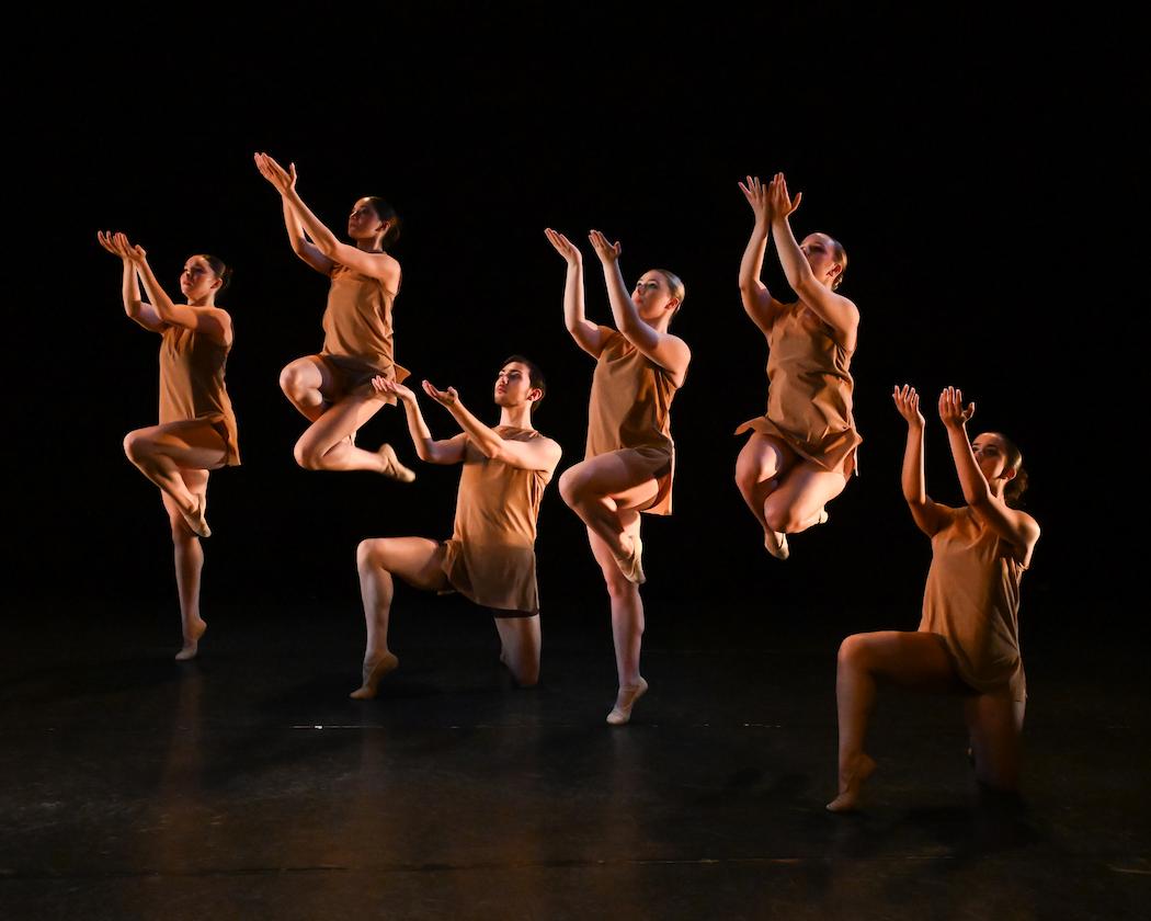 Stage image from an Emerging Choreographers Showcase showing a group of dancers arranged in a dynamic formation. Each dancer wears a simple, warm-toned costume and holds a stylized pose—some kneeling, others lifted or balanced—with arms raised and angled to create strong geometric lines. The performance takes place on a dark stage with focused lighting that highlights the dancers’ forms and synchronized composition.
