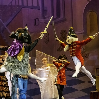 Stage scene from The Nutcracker showing performers in elaborate costumes, including a horned creature with shaggy textures and a toy soldier in a red uniform and tall hat, both mid-leap while fencing with swords. Additional dancers in bright costumes appear behind them, with a stylized backdrop of interior architecture in pink and purple tones.