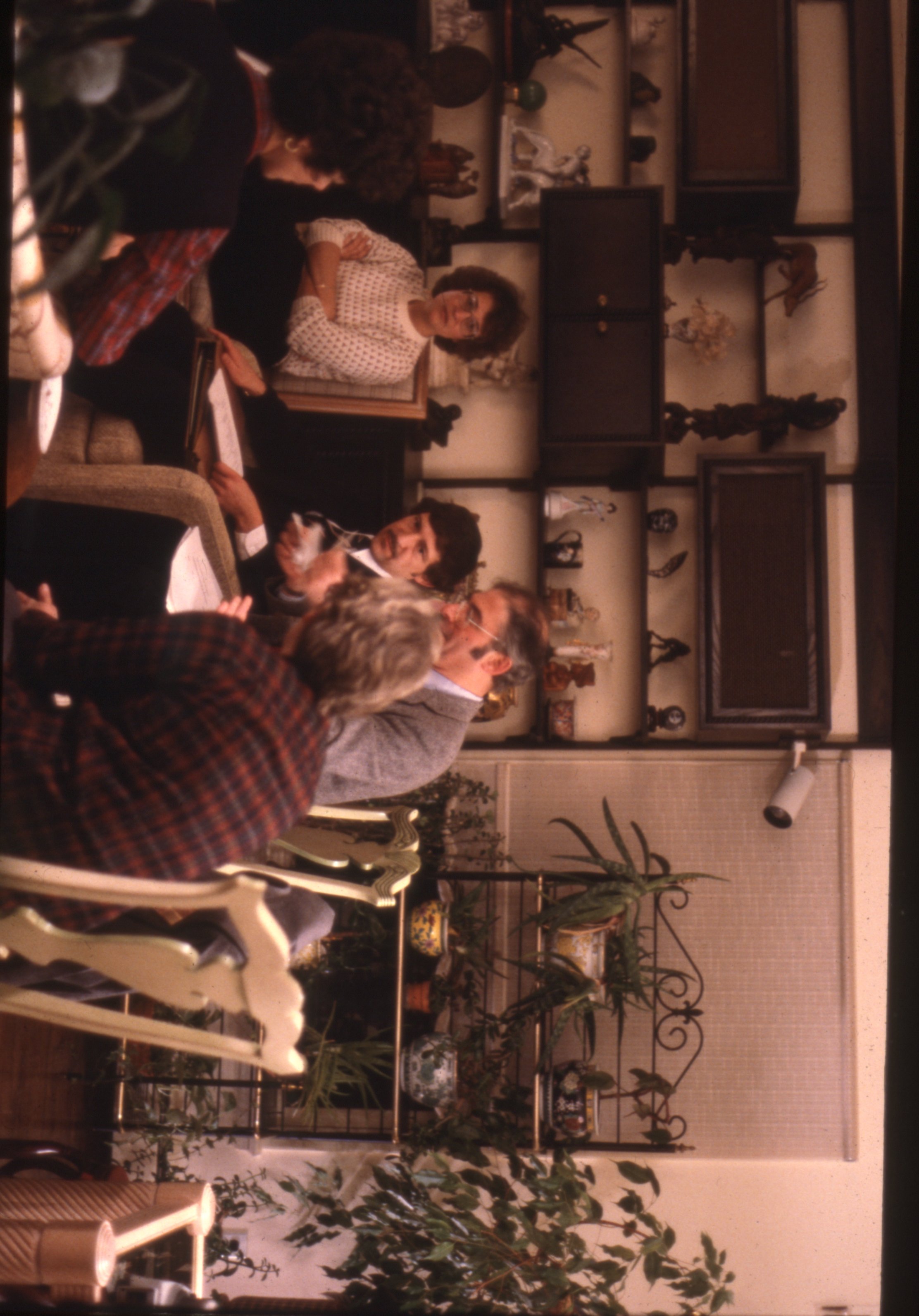 Archival photograph of a small group gathered in a residential living room for a meeting, seated among household furnishings, decorative shelves, and indoor plants.