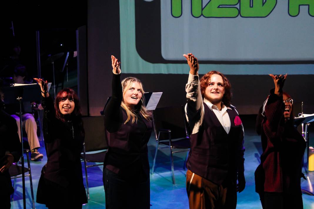 Stage image from a Theatre New Works Showcase featuring four performers standing in a row, each extending one hand upward in a unified gesture. The performers wear dark clothing, and music stands are visible nearby, suggesting a staged reading or presentation. The background includes a large projection screen with partial text, and the stage lighting casts cool tones across the scene.